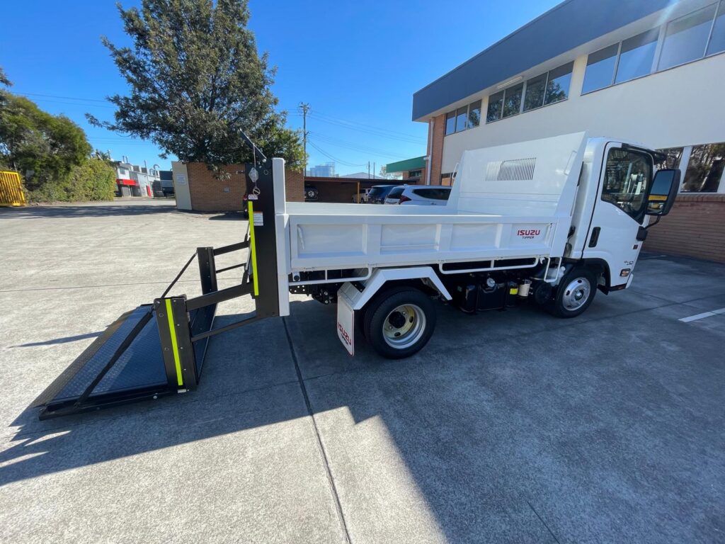 Isuzu flatbed truck with lowered hydraulic tailgate/liftgate ramp, parked in an outdoor lot.